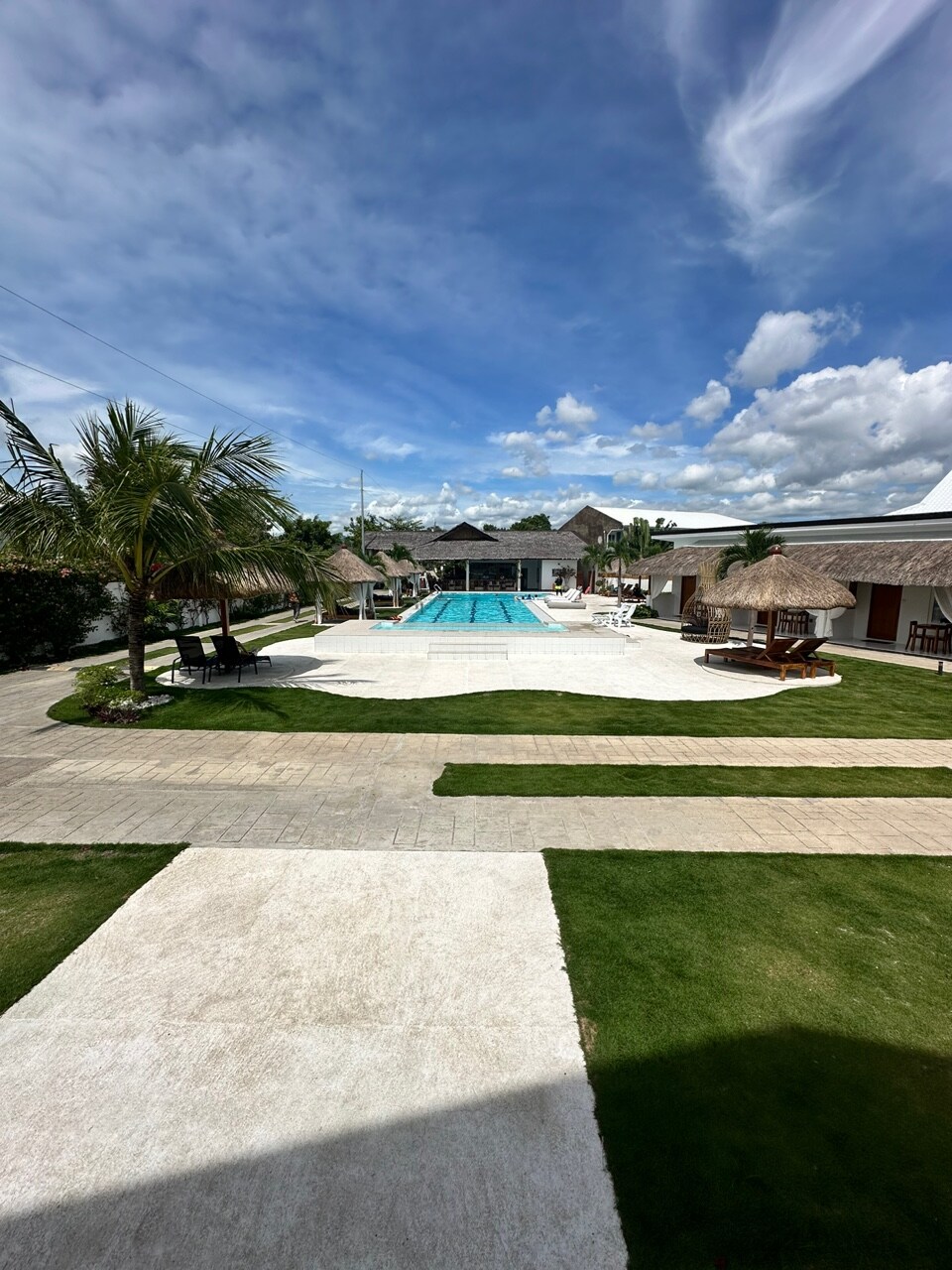 The outdoor area features a spacious pool surrounded by lounge chairs and palm trees. A pathway leads to the pool, while grassy areas enhance the inviting environment. Thatched-roof structures provide shaded seating, and bright blue skies are visible overhead, adding to the overall charm.