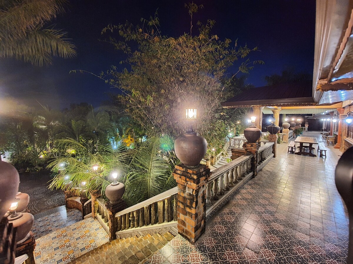 A well-lit pathway is lined with lanterns, guiding the way down a staircase adorned with decorative urns. Lush greenery and palm trees create a serene backdrop, while a dining area with a long table is visible in the distance, enhancing the inviting atmosphere of the outdoor space.