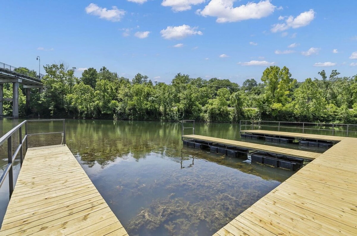 A wooden dock extends over calm waters, bordered by lush greenery on either side. The clear blue sky is dotted with clouds, reflecting on the surface of the water. A bridge is visible in the background, adding to the serene outdoor setting.