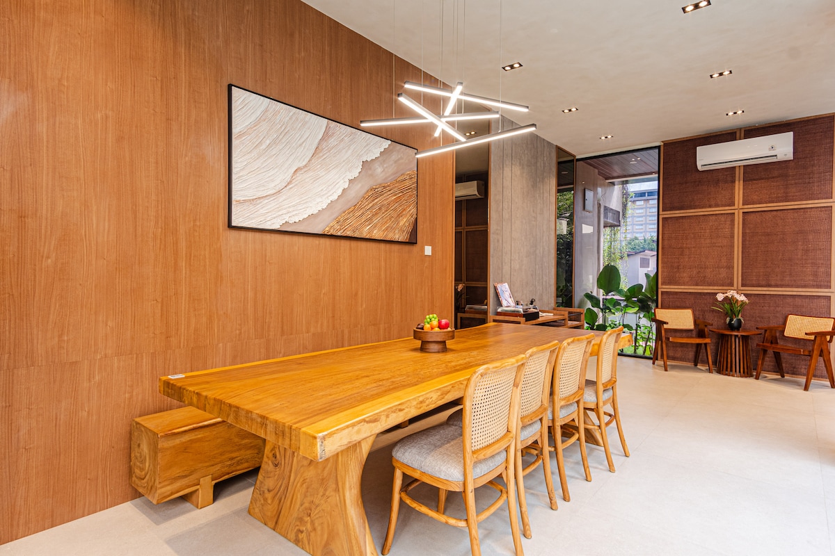 A spacious dining area features a large wooden table surrounded by several light-colored chairs. A striking light fixture hangs above, while a piece of art is displayed on the wall. Natural light enters through large windows, complementing the warm wooden accents.