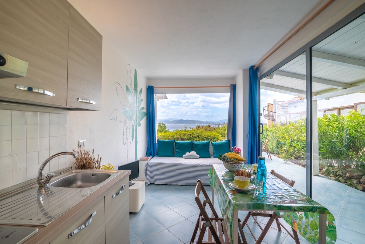 A functional living area is displayed, featuring a kitchenette with ample counter space and a dining table covered with a decorative cloth. A comfortable seating area with colorful cushions is visible through large glass doors, leading to an outdoor terrace with verdant landscaping.