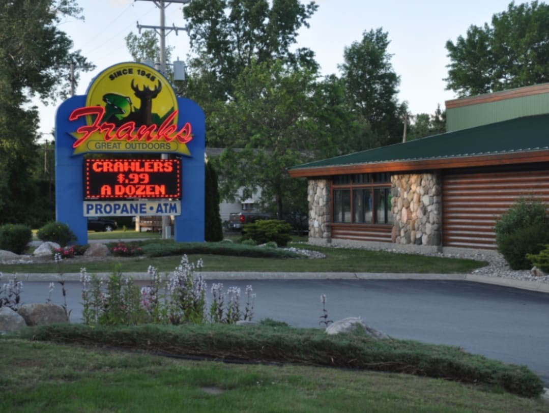 A stone-accented building features a large, colorful sign for Frank's Great Outdoors. The sign displays prices for fishing supplies and promotes the availability of propane and an ATM. Lush trees and landscaped plants surround the structure, contributing to a welcoming outdoor environment.