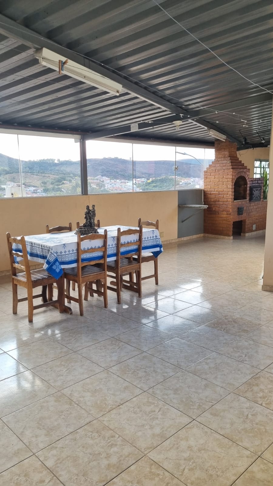 A spacious terrace features a wooden dining table set for six, complemented by a near view of a brick barbecue grill. Large windows offer natural light and scenic mountain views, while tiled flooring enhances the open feel of the area.