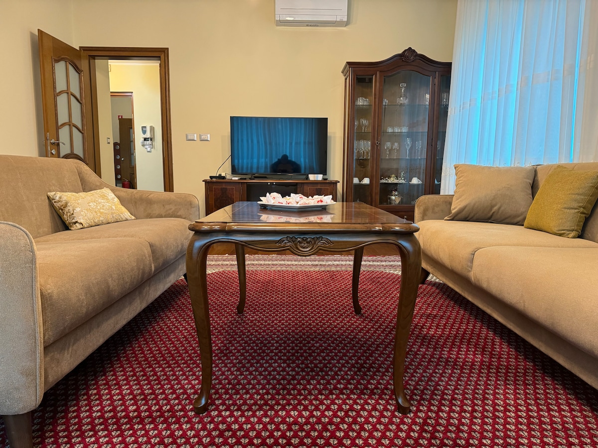 A cozy living area is presented with two beige sofas arranged around a wooden coffee table. The rich red carpet adds warmth to the space. A television is positioned on a cabinet, with natural light filtering through light curtains from a nearby window.