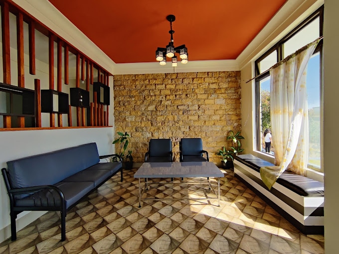 A spacious seating area features a large window allowing natural light to fill the room. Dark blue sofas face two black chairs, arranged around a sleek coffee table. Decorative plants are positioned near the window, enhancing the relaxed atmosphere.