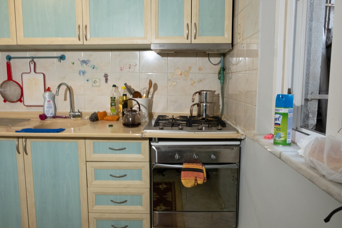 The kitchen features light wooden cabinetry with pastel blue accents. A gas stove is positioned next to a sink, with utensils and cleaning supplies readily available. Natural light enters through a nearby window, illuminating the space, which offers essential amenities for meal preparation.