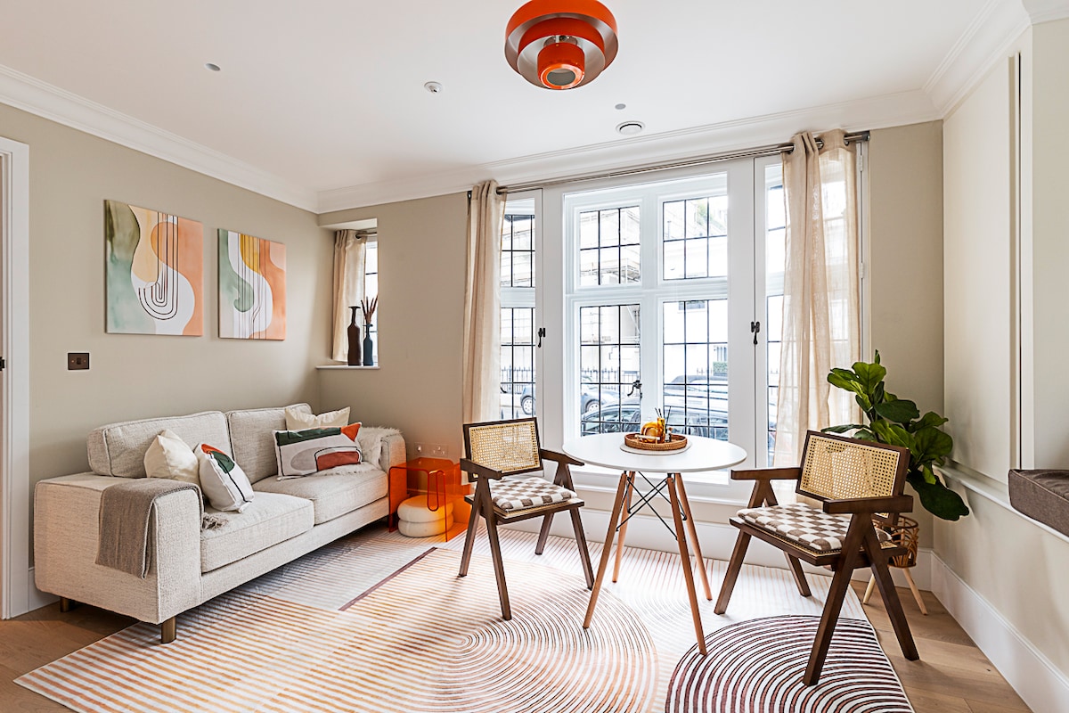 A light-filled living area features a comfortable sofa with decorative cushions, accompanied by two wooden chairs. A round table sits at the center, enhanced by a bowl of fresh fruit. Large windows allow natural light to illuminate the soft, neutral tones of the space.