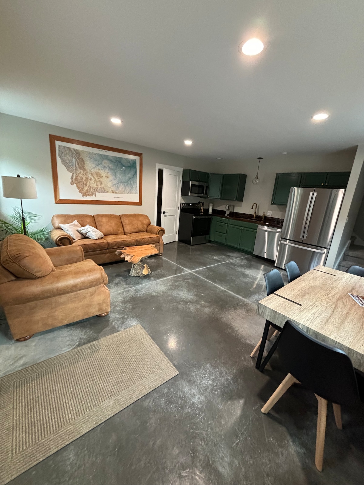 A welcoming living space features a brown sofa paired with a wooden side table. A dining area includes a natural wood table surrounded by black chairs. The kitchen is equipped with modern appliances, and a colorful artwork hangs on the wall, enhancing the ambiance.