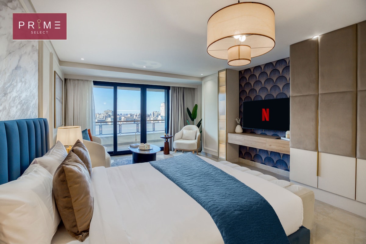 A modern bedroom features a spacious bed adorned with crisp white linens and accent pillows. Large windows offer views of the river and cityscape. A comfortable armchair is positioned near the window, and a wall-mounted TV is visible across the room, creating a relaxing atmosphere.