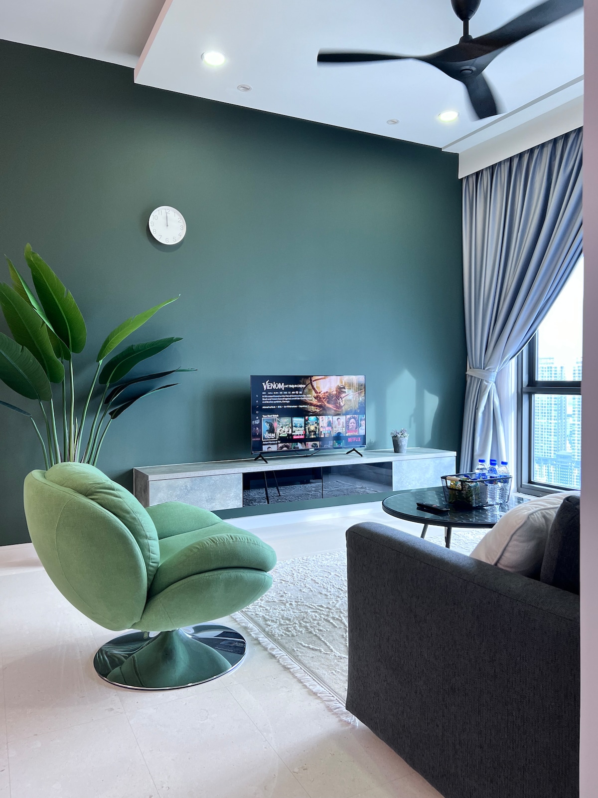 A modern living area features a plush green swivel chair and a gray sofa, both positioned around a sleek coffee table. A wall-mounted TV displays content, while large windows dressed in light curtains enhance the space with natural light and city views.