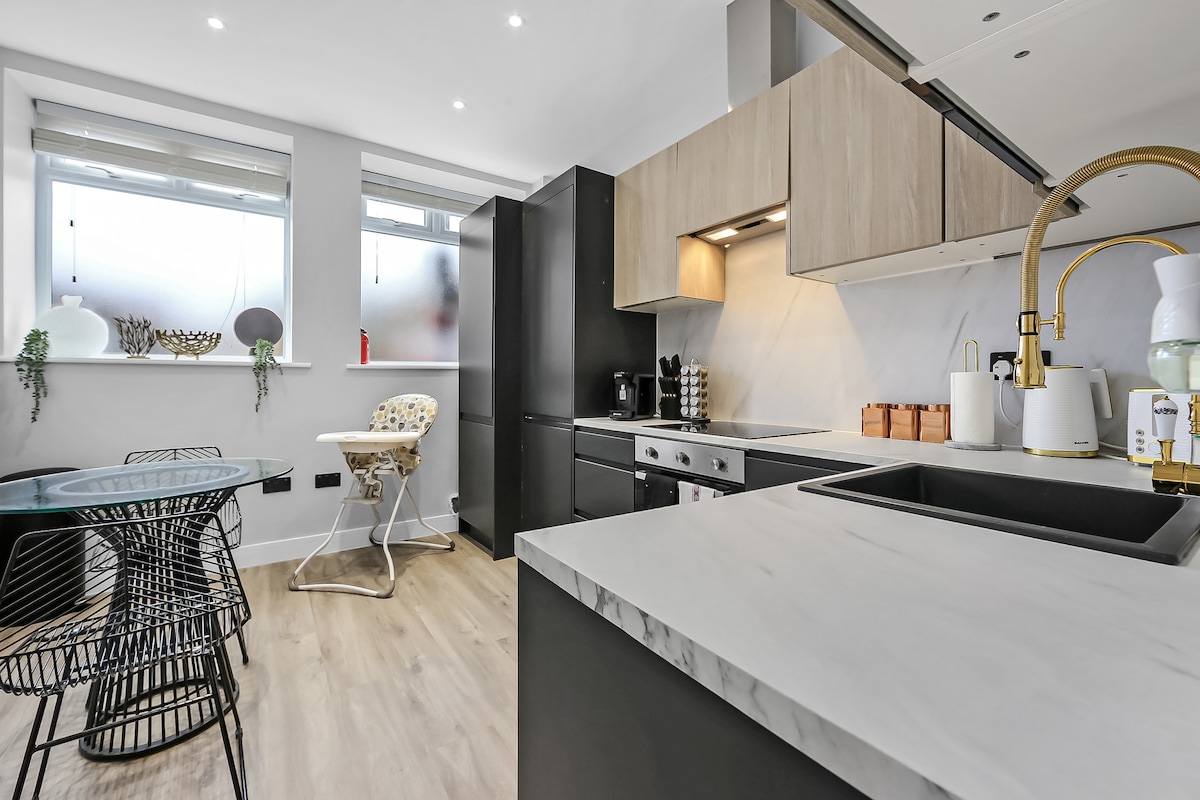 A modern kitchen is displayed with sleek cabinetry and a marble countertop. An elegant gold faucet complements the simple sink. A dining table with stylish black chairs is positioned beside large windows that allow natural light to fill the space.