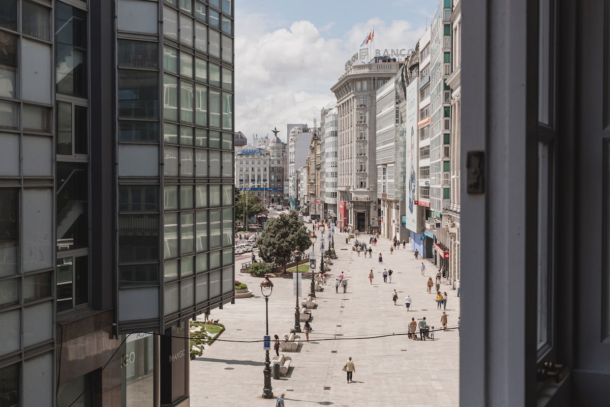 A view from the window showcases a bustling urban street lined with shops and modern buildings. Pedestrians can be seen walking along the broad walkway. Lush trees and streetlamps are positioned throughout the area, creating a vibrant atmosphere in the heart of the city.