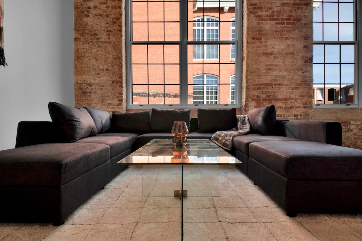 A spacious living area features a large sectional sofa in a dark fabric, complemented by a glass coffee table. Natural light floods in through tall windows, highlighting the exposed brick walls and creating a modern, airy feel.