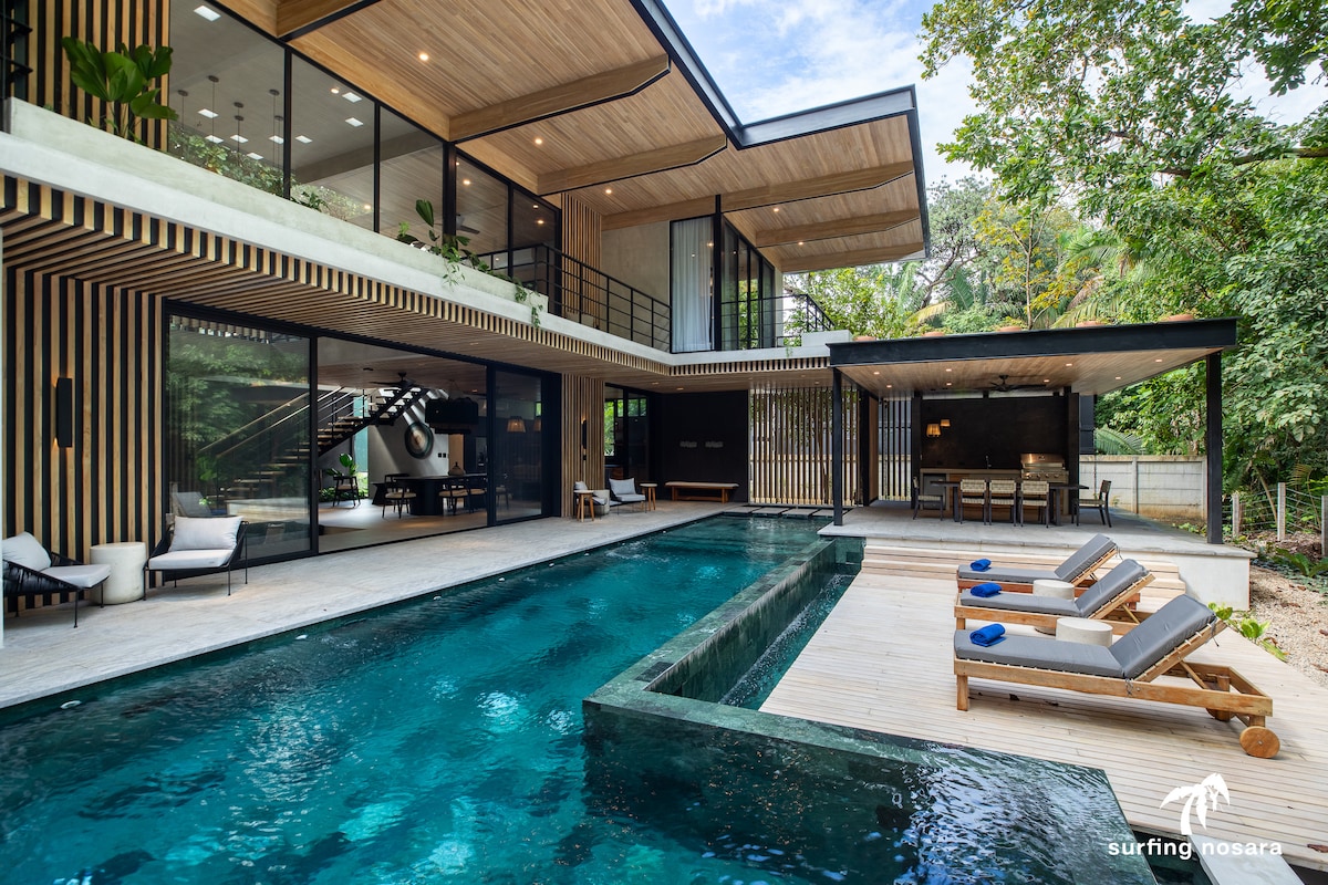 The villa features a striking infinity pool set against a backdrop of lush jungle, with modern lounge chairs positioned for relaxation. Large glass walls create an openness, allowing natural light to fill the indoor spaces, which display a blend of wood and contemporary design.