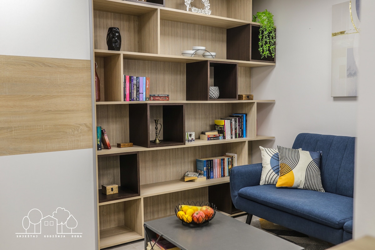 A modern shelving unit displays books and decorative items, providing a blend of storage and style. A comfortable blue sofa is positioned in front, adorned with colorful cushions. A coffee table sets the stage for a bowl of fruit, adding a touch of warmth.