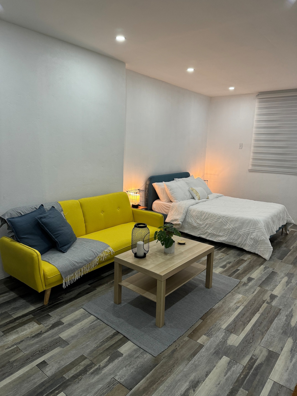 A comfortable living area features a bright yellow sofa accompanied by decorative pillows. A small wooden table is placed in front, with a potted plant and lantern. A cozy sleeping space includes a spacious bed with soft bedding, located against a well-lit wall with subtle textures.