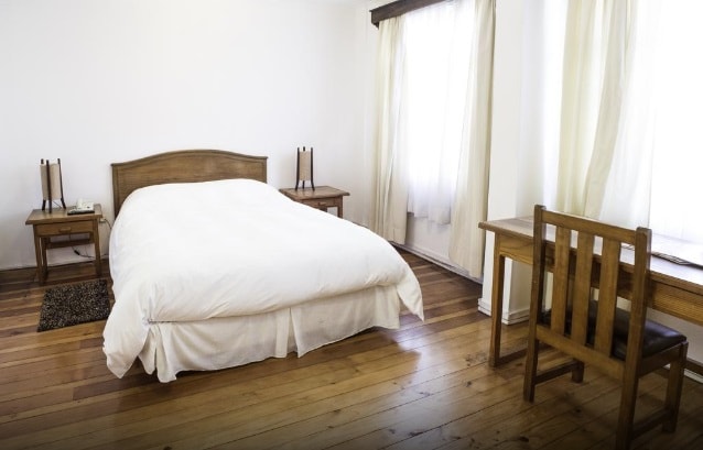 A spacious bedroom features a large bed with a crisp white duvet and wooden accents. Two bedside tables are situated beside the bed, each with a lamp. A wooden desk and chair are positioned nearby, with natural light streaming through the sheer curtains of a window.