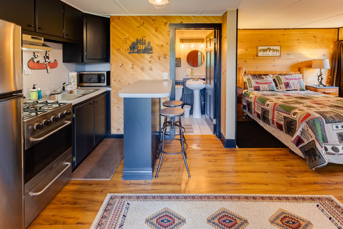 A cozy studio layout is shown, featuring a full kitchen with stainless steel appliances and a breakfast bar with two stools. The sleeping area has a queen bed adorned with a patterned blanket. Warm wood paneling complements the space, creating a rustic atmosphere.