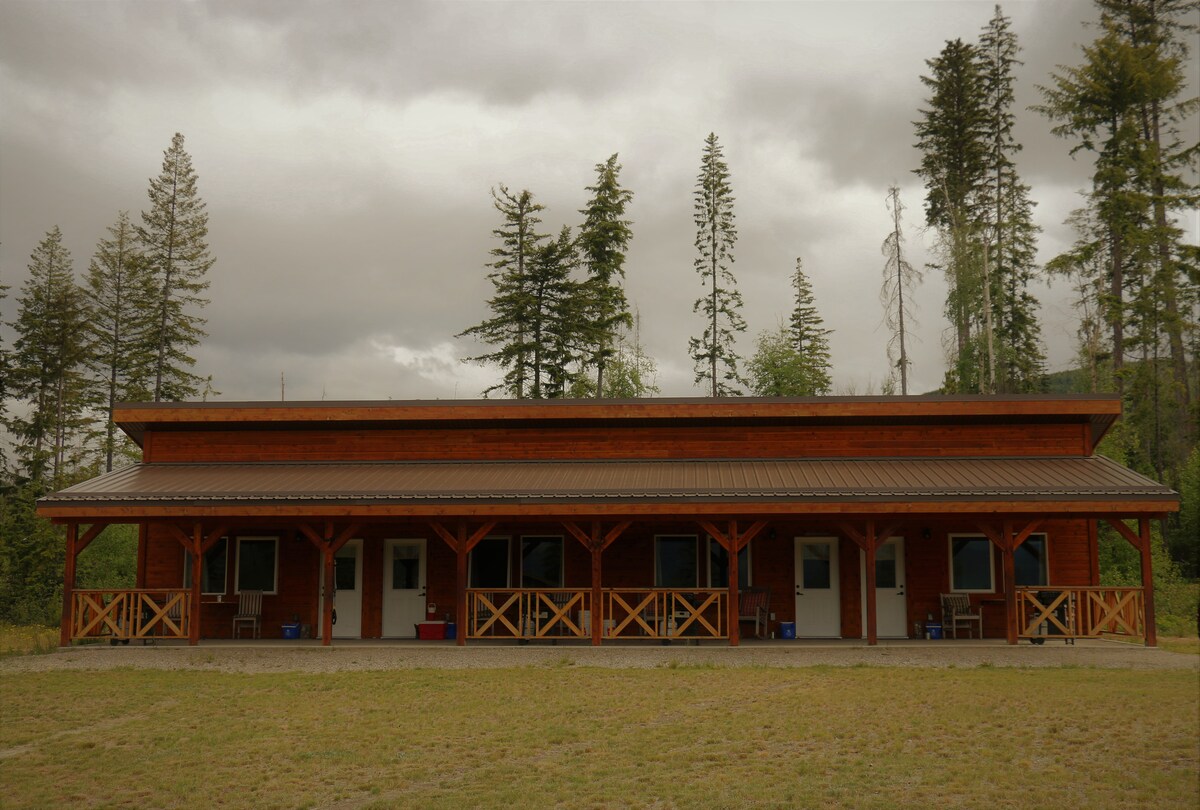 A multi-unit cabin structure is set against a backdrop of tall trees. The exterior features a wooden façade with a sheltered porch, providing access to individual entrances. The surrounding area consists of an open grassy space, enhancing the cabin's natural setting.