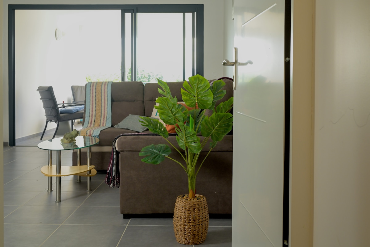 The image captures a view from the entrance, showcasing a modern living area. A comfortable brown sofa is adorned with a colorful throw, complemented by a glass coffee table. A potted plant adds a touch of greenery, while a dining table is visible in the background.