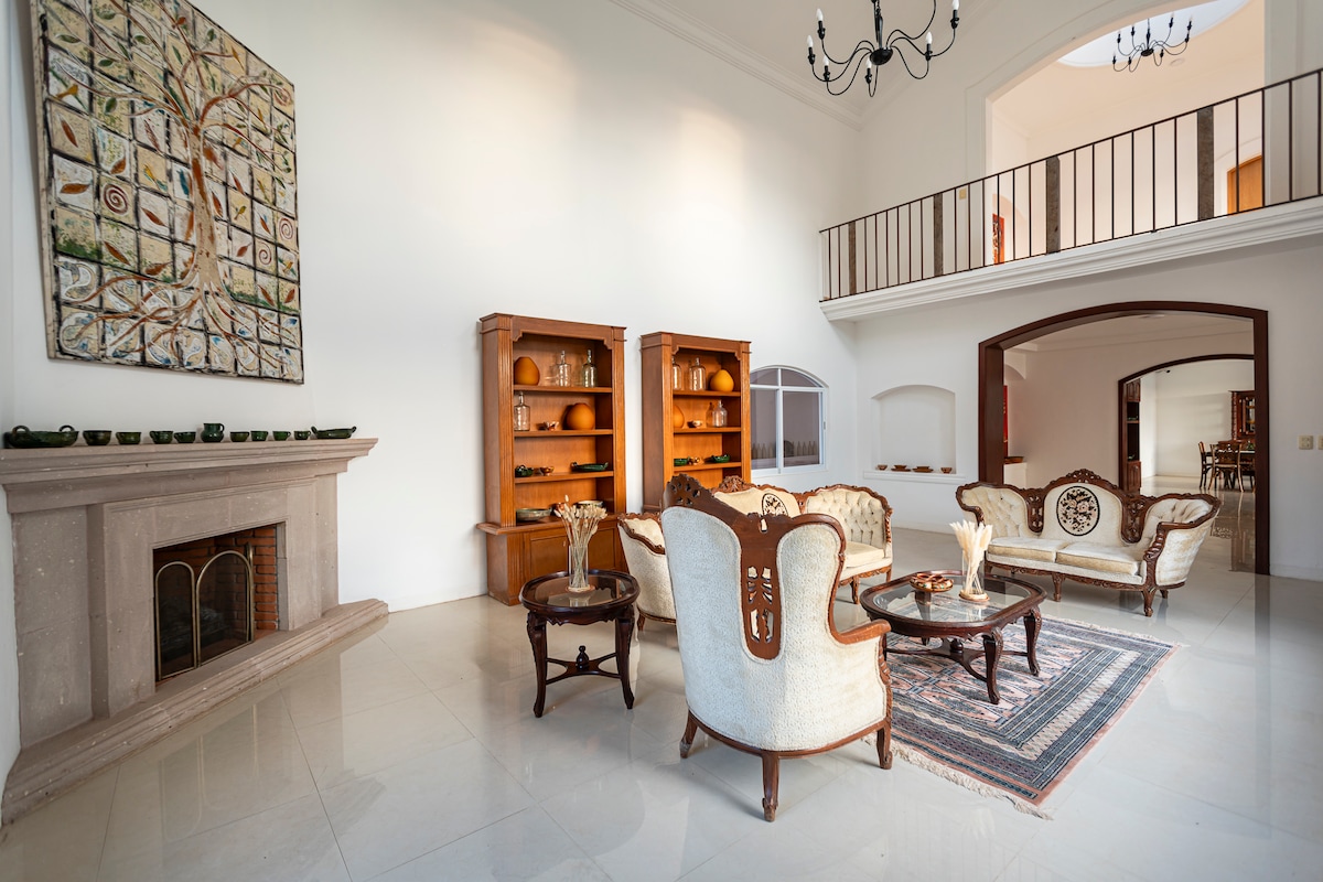 A spacious living area features a vintage-style seating arrangement centered around a low table. Two wooden bookshelves display decorative items. A large piece of wall art provides a focal point above a stylish fireplace, with natural light filtering in through the arched entryway.