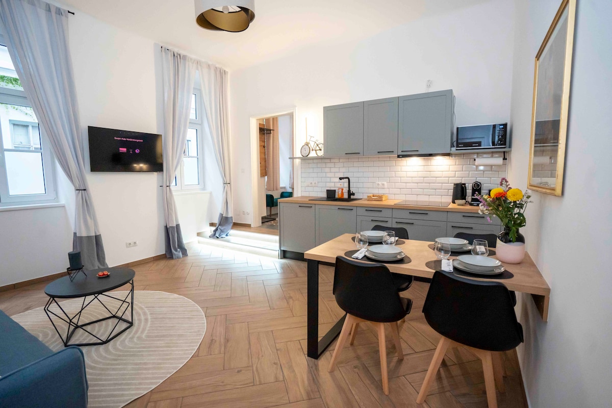 A modern living area is shown, featuring a dining table with two black chairs next to a well-equipped kitchen. Soft curtains hang by the windows, allowing natural light to illuminate the space. A cozy seating area includes a light blue couch and a round coffee table.