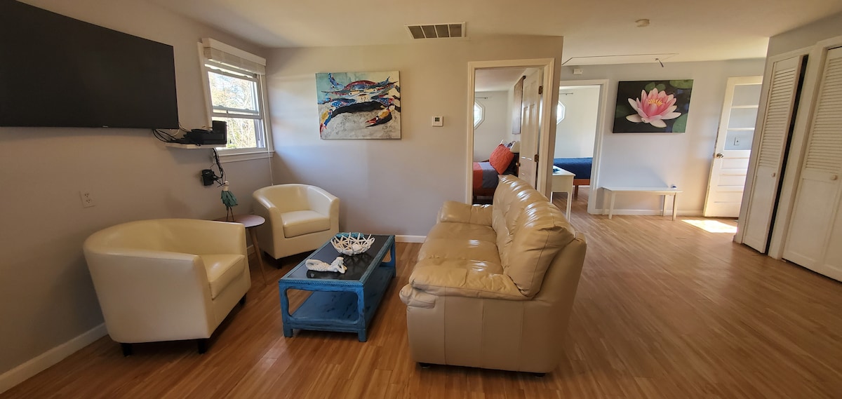A bright living area features a comfortable leather sofa and two white armchairs arranged around a blue coffee table. Artwork adorns the walls, and a wall-mounted TV is visible. Natural light enters through the windows, illuminating the warm wood flooring.