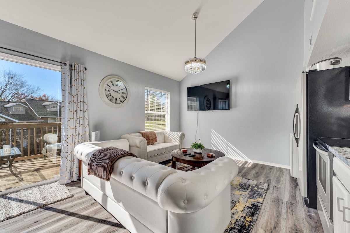 A bright living room features a large window allowing natural light to fill the space. A modern chandelier hangs above a white leather sectional sofa and a coffee table. A flat-screen TV is mounted on the wall, and a balcony is visible through the sliding glass door.