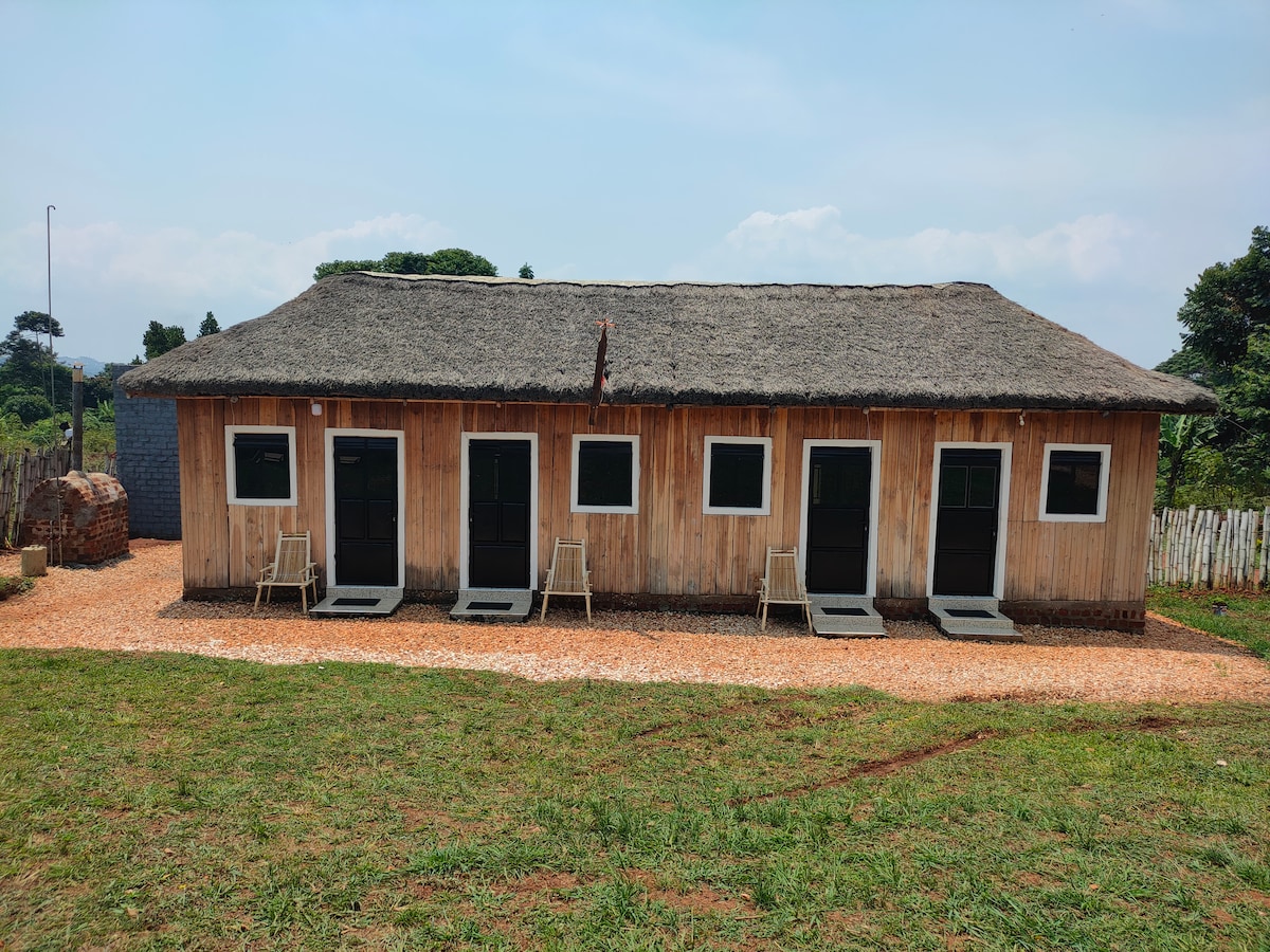 A charming thatched cottage suite is presented, featuring a wooden exterior and multiple dark doors. Simple seating is placed outside each door on a gravel path. The surrounding green landscape and blue sky enhance the tranquil outdoor setting.