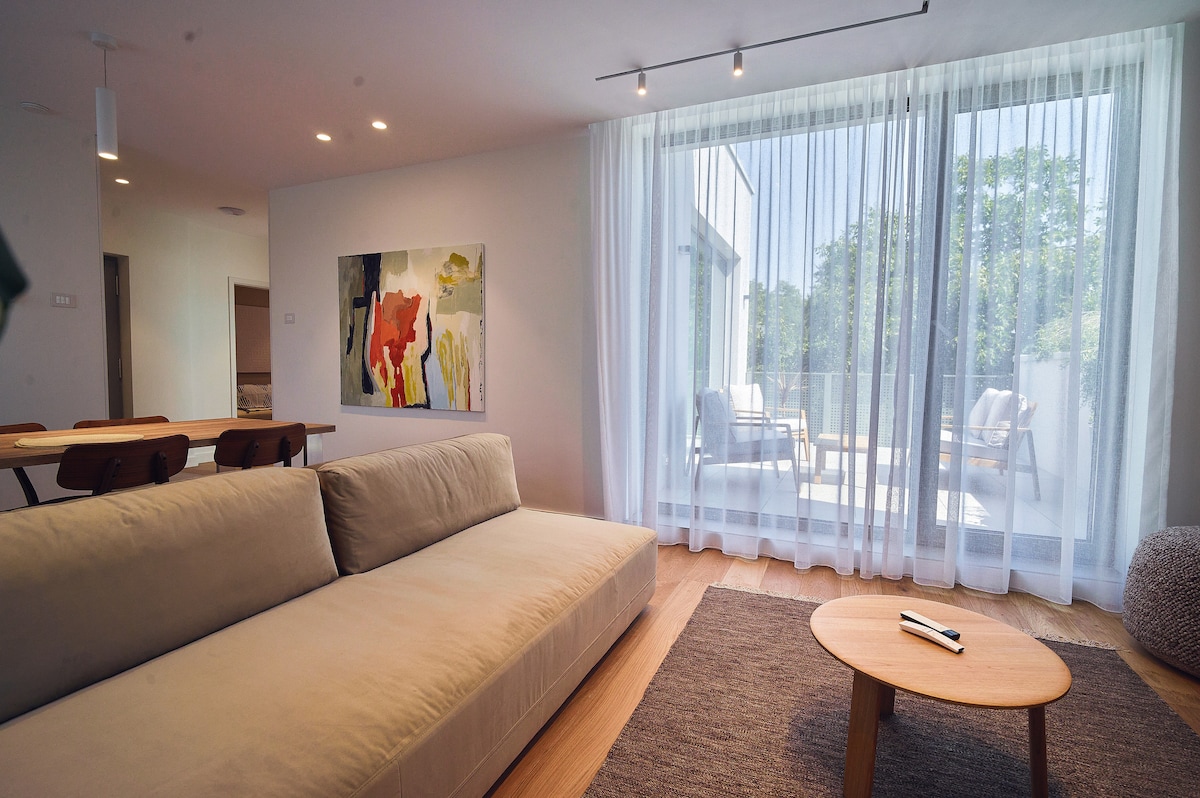 A spacious living area is highlighted by a light-colored sofa, complemented by a wooden coffee table. Floor-to-ceiling sheer curtains allow natural light to illuminate the space, while a dining area is visible in the background, featuring a wooden table and chairs. A balcony can be seen through the curtains.