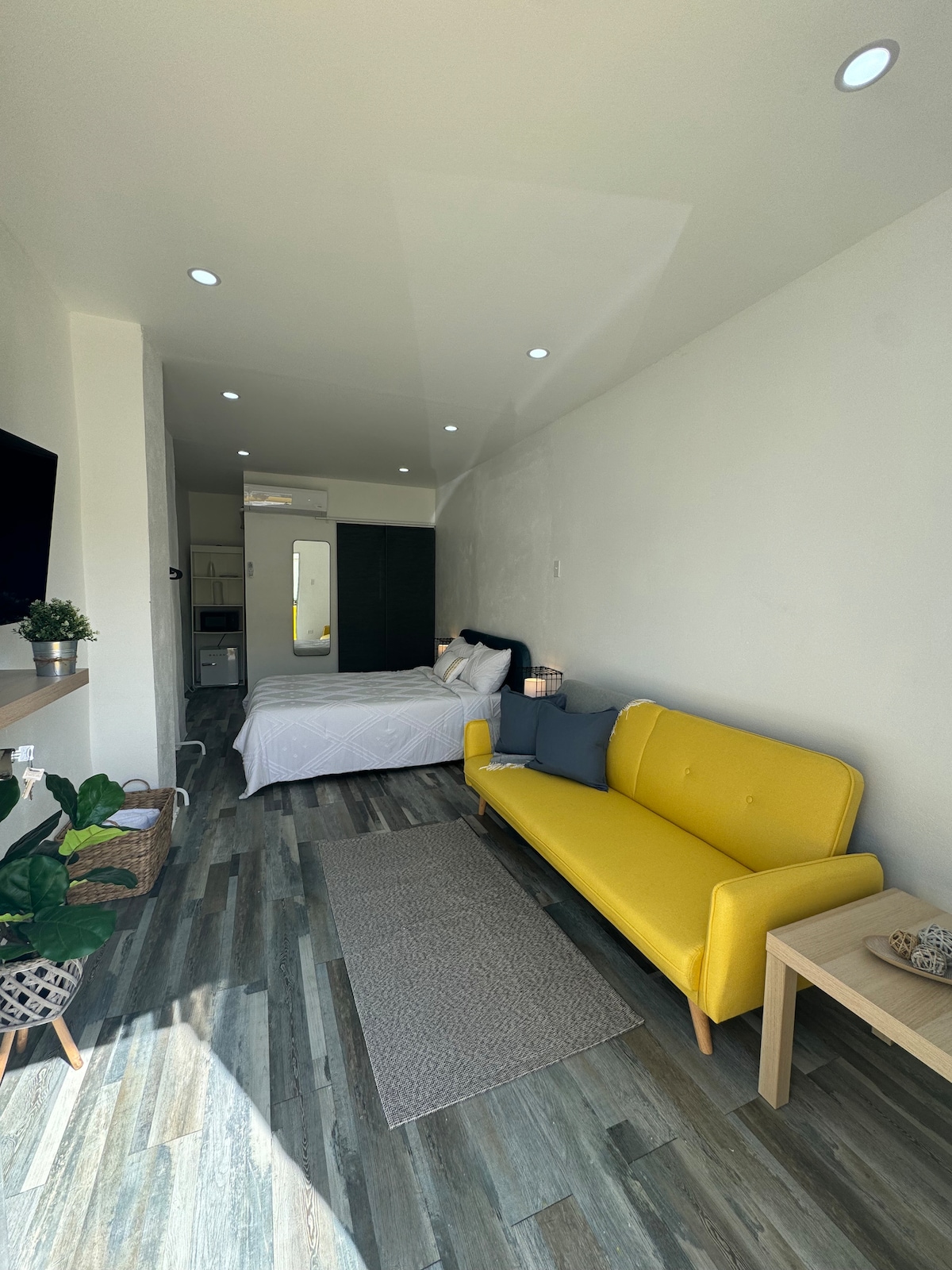 A studio apartment features a bright yellow sofa positioned in the foreground, complemented by a light wooden side table. A spacious bed is visible in the background, and soft lighting is provided by recessed ceiling lights. Plants add a touch of greenery to the decor.