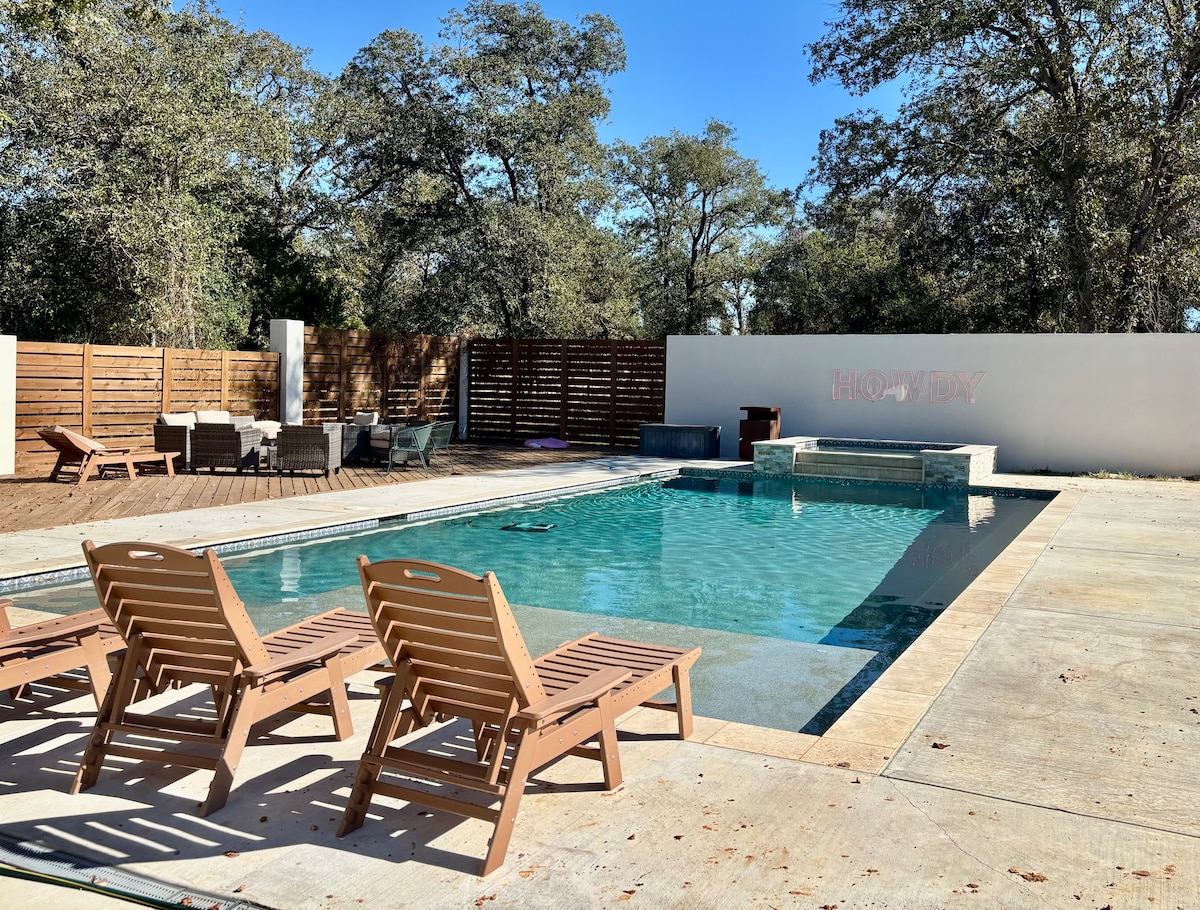 A well-maintained swimming pool is featured, surrounded by wooden lounge chairs arranged on a spacious patio. A wooden fence provides privacy, while shaded seating areas can be seen in the background. The pool's clear water reflects the sunlight on a bright day.