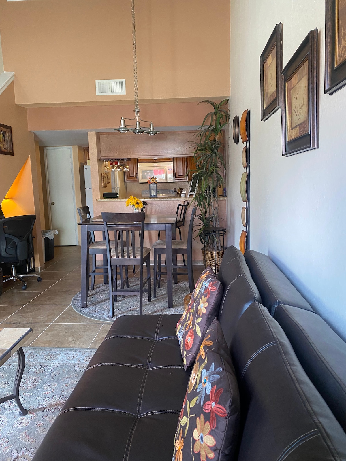 A comfortable living area features a dark leather sofa adorned with decorative pillows. A dining set with tall chairs is positioned near a well-equipped kitchen, with warm tones enhancing the inviting space. A small table sits in front of the sofa, completing the layout.