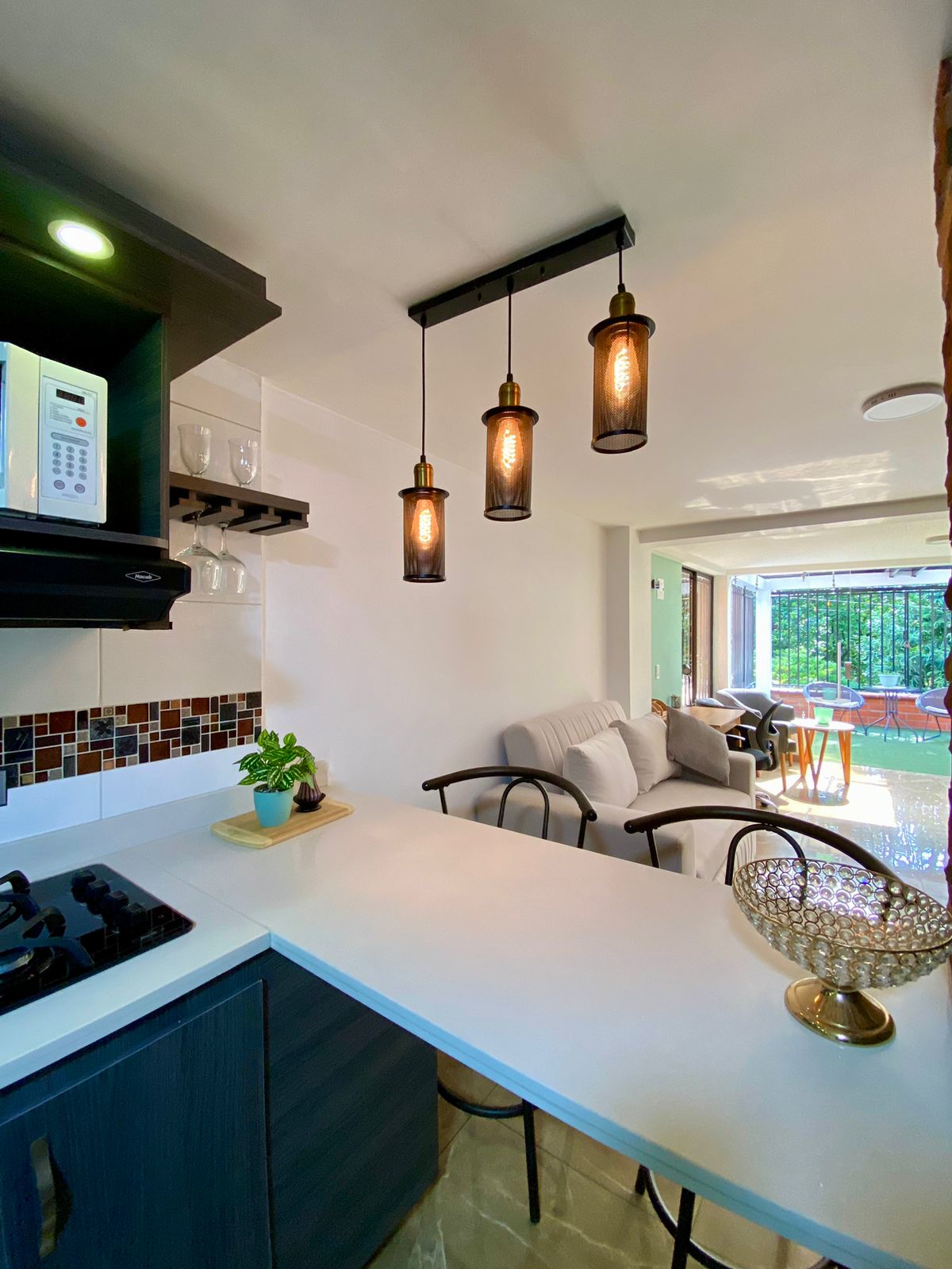 An open kitchen area features a modern design with dark cabinetry and a white countertop. Three pendant lights hang above, illuminating the space. A small plant adds a touch of greenery, while a living area with comfortable seating is visible through sliding glass doors.