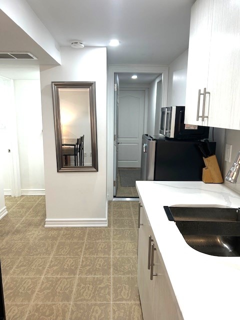 The kitchen area features modern cabinetry and a sleek countertop with a sink. A mirror reflects the space, while a hallway leads to a door in the background. A fridge and microwave are visible, contributing to the functional design.