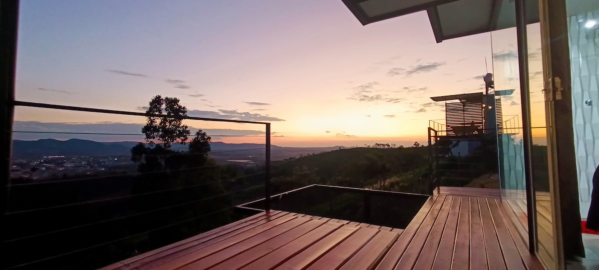A wooden deck extends outward, offering a panoramic view of the surrounding mountains at sunset. The sky displays warm hues of orange and purple. A sleek structure is visible to the right, framed by large glass windows, enhancing the connection with nature.