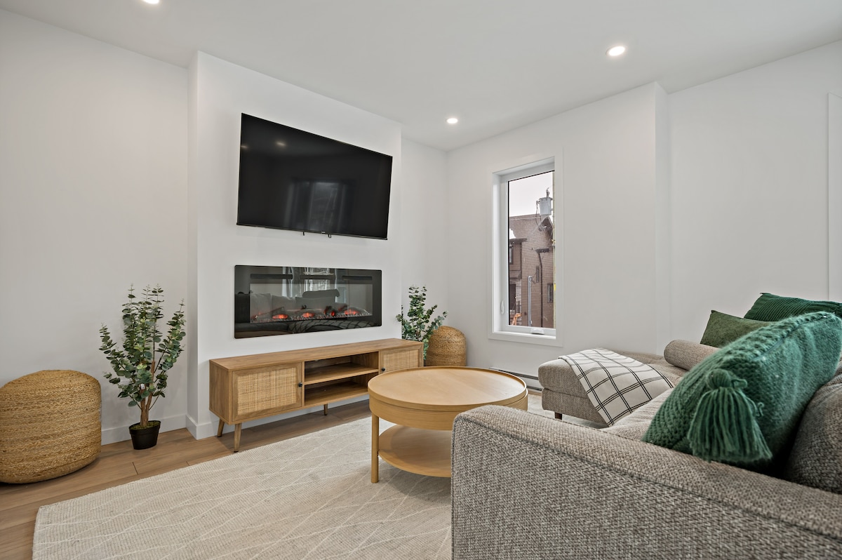 A modern living room is highlighted by a wall-mounted television and a sleek electric fireplace below. A round coffee table occupies the center, surrounded by a comfortable sofa adorned with green and patterned cushions. Natural light filters in through a nearby window, illuminating potted plants.