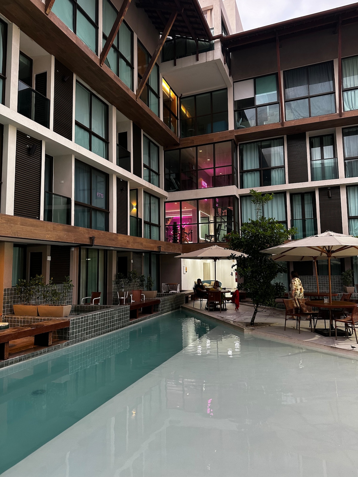 The image captures a serene pool area bordered by modern architecture. Wooden seating and tables are positioned near the pool, alongside vibrant greenery. Large windows reflect warm lighting from the upper levels, creating a welcoming atmosphere within the condominium.