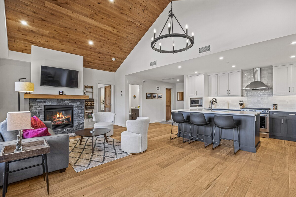 An open-plan living area features vaulted wood ceilings and large windows allowing natural light. A modern kitchen with a large island and seating faces a cozy sitting area, which includes a fireplace and comfortable chairs. Neutral tones with pops of color add to the inviting space.