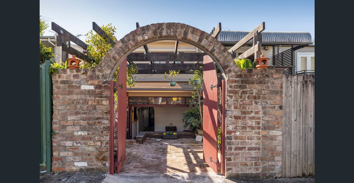 A charming entrance is framed by a brick archway leading into a private courtyard. Red double doors are open, revealing a paved area lined with greenery and potted plants. Above, a wooden pergola provides partial shade, enhancing the inviting outdoor space.