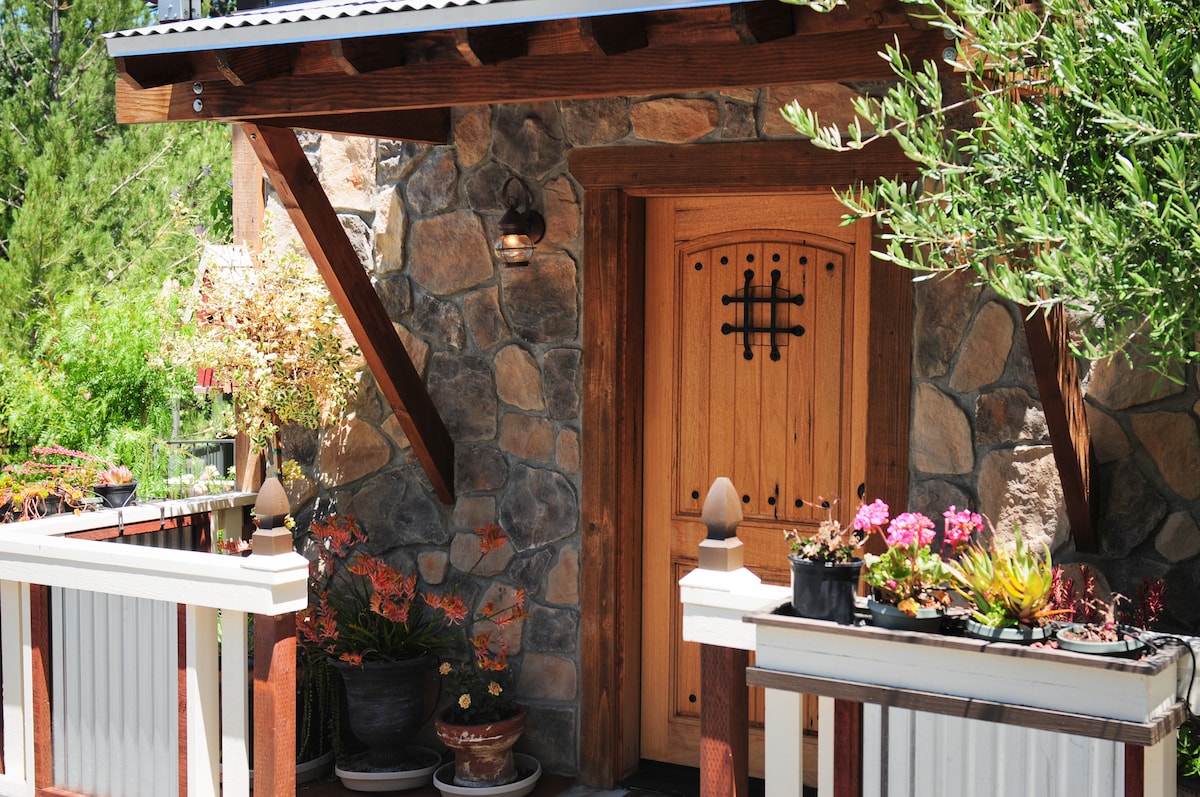 A rustic entrance features a wooden door with a decorative wrought iron grate, surrounded by natural stone walls. Potted plants add a touch of green and color to the porch area, enhancing the inviting exterior of the building.