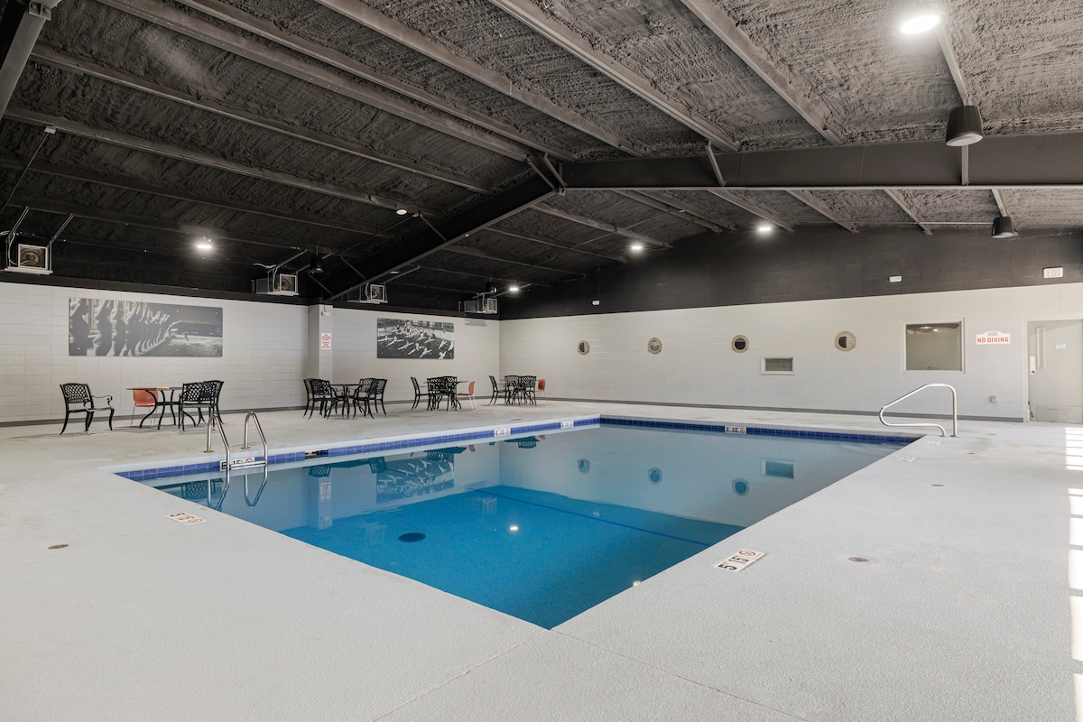 An indoor pool area is featured, with a spacious pool surrounded by a white deck. Several black metal chairs and tables are arranged nearby. Soft lighting illuminates the high ceiling, creating a welcoming environment for relaxation.