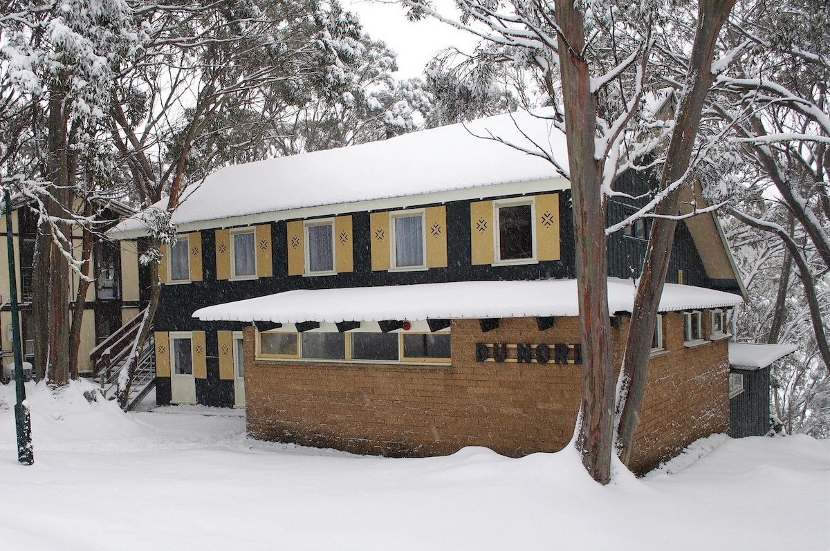 The exterior of a cozy lodge is showcased, surrounded by tall trees blanketed in snow. The rustic building features a two-story structure with large windows, framed in dark wood. A sign reading 'Du Nord' is visible on the snow-covered front wall.