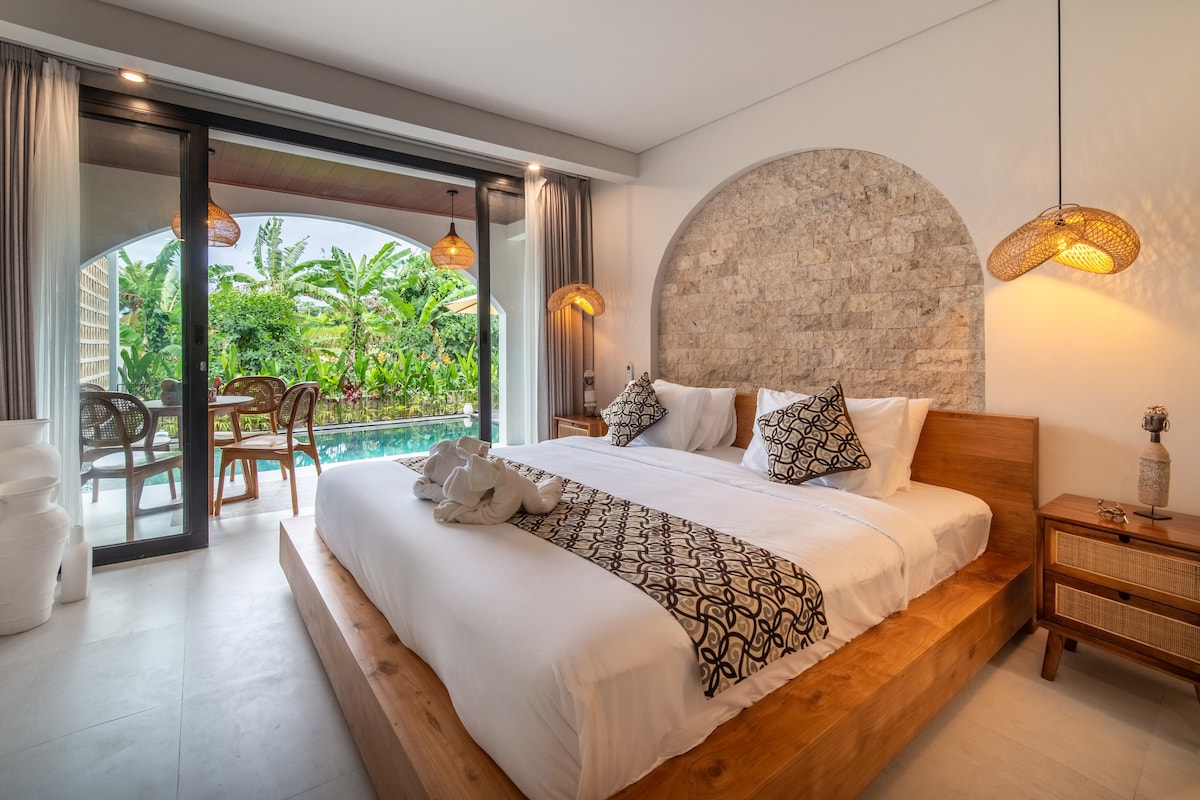 A spacious bedroom features a king-size bed adorned with decorative pillows. Large glass doors open to a private terrace with views of lush greenery. Soft lighting from woven pendant lamps enhances the serene environment, complemented by natural stone accents on the walls.