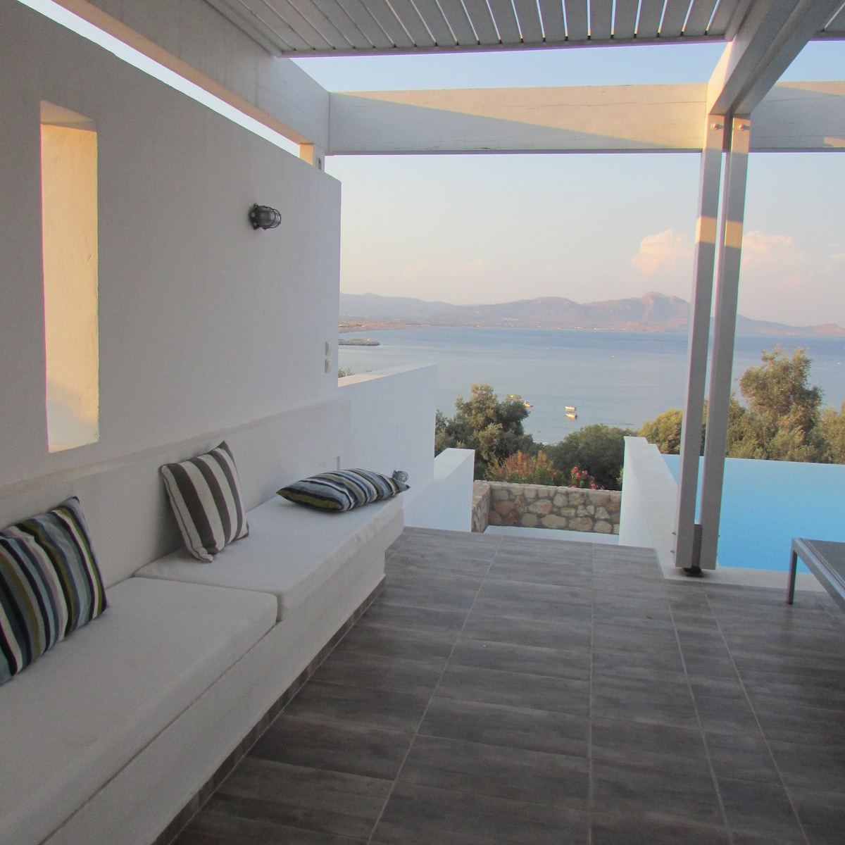 An outdoor terrace is showcased, featuring a built-in cushioned seating area with striped pillows. A stunning view of the Aegean Sea and distant mountains can be seen through the open space, along with a glimpse of the infinity pool reflecting the sky.