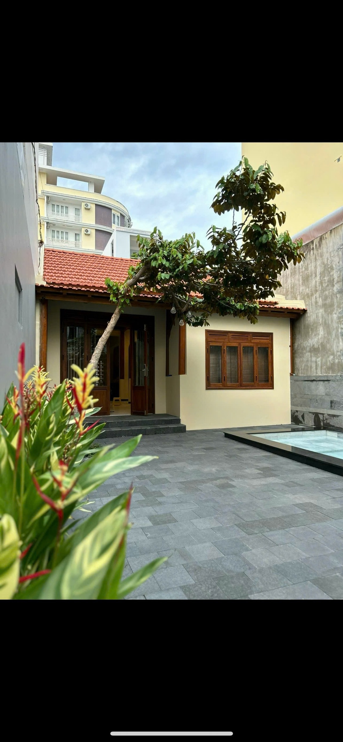 An exterior view of the villa is presented, featuring a single-story structure with a tiled roof and wooden accents. The entrance is framed by lush, tropical plants, adding a touch of greenery. A tranquil pool can be seen beside the house, complementing the peaceful environment.