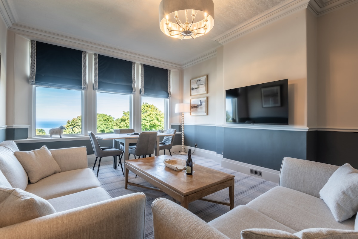 The living area is bright and airy, featuring large windows that offer views of the landscape. A comfortable seating arrangement with light-colored sofas surrounds a wooden coffee table. A dining area with modern chairs is visible, while a television is mounted on the wall.
