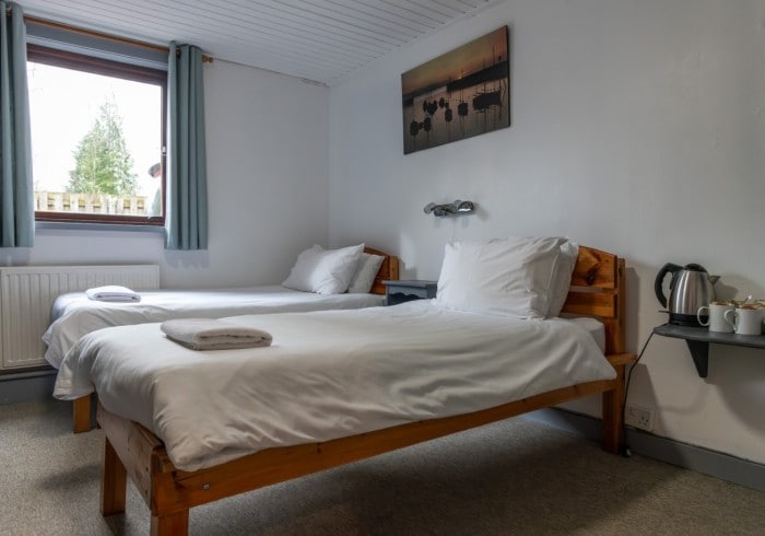 A simple guest room is presented with two wooden framed beds, each topped with white linens and a folded towel. A small bedside table is situated between the beds. A window with light blue curtains allows natural light to enter, and a wall-mounted picture adds visual interest.