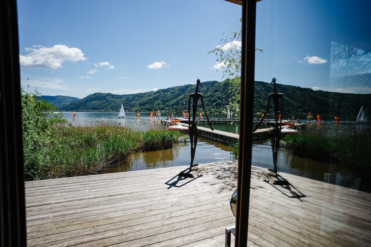 A serene view of the lake is visible through the glass doors, with lush greenery framing the shoreline. Wooden decking leads to a small pier, where sculptural figures stand. Sailboats can be seen gliding across the water, under a bright blue sky dotted with clouds.