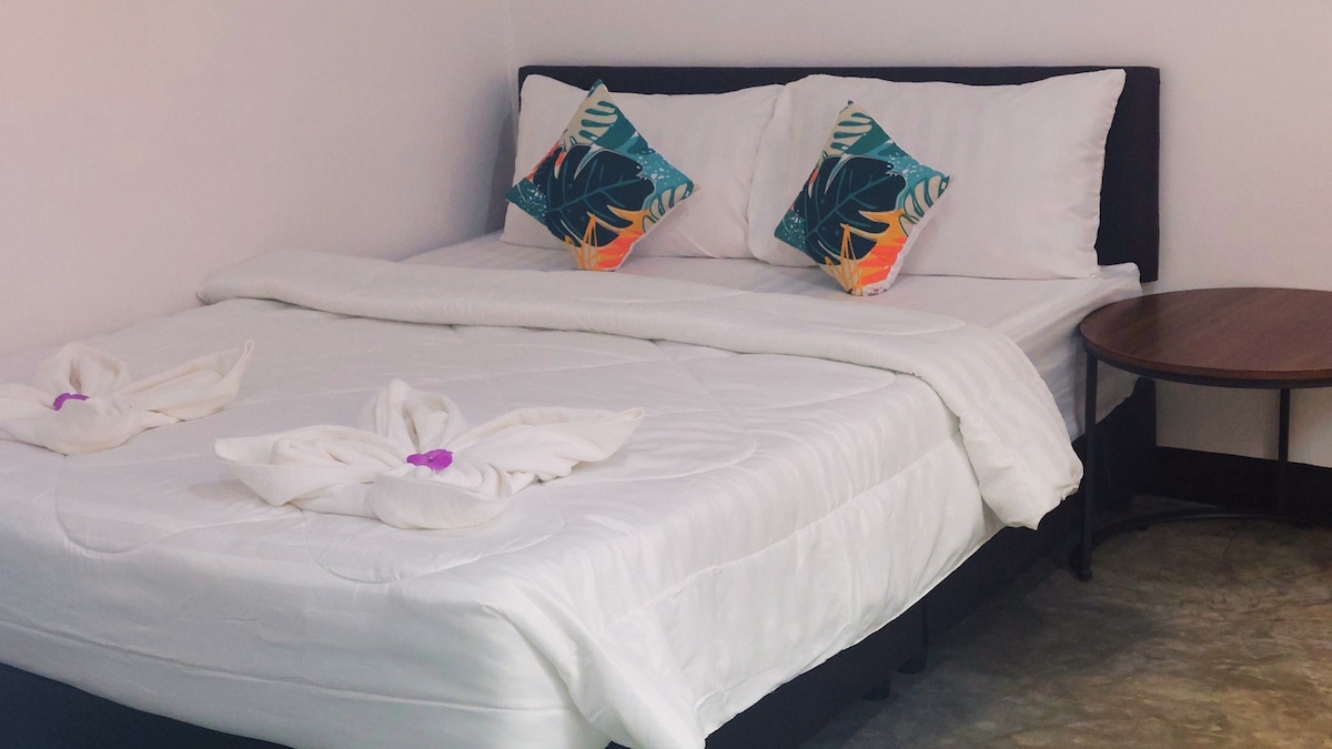 A comfortable sleeping area is furnished with a spacious bed adorned with white linens. Two decorative cushions with tropical patterns are placed at the head of the bed. A small side table is positioned beside the bed, contributing to a cozy atmosphere.